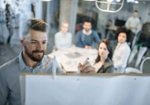 German course in a company. The teacher is writing on a flip-chart, the course participants are sitting around a table.