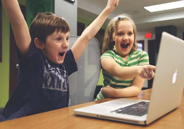 Two kids are learning German online and are sitting in front of a computer, cheering. Photographer > getty images